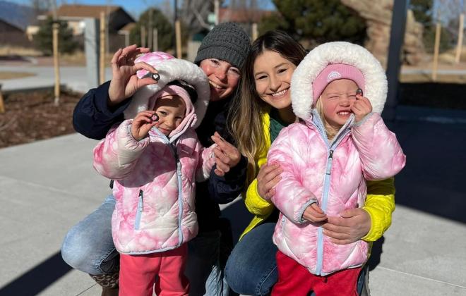 two little grils in pink coats with two women. The little girls are showing off tiny pink hearts.