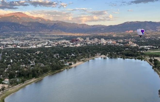 An aerial view of Prospect Lake and Memorial Park on a sunny morning. A hot air balloon floats over the lake and Pikes Peak is visible in the background.